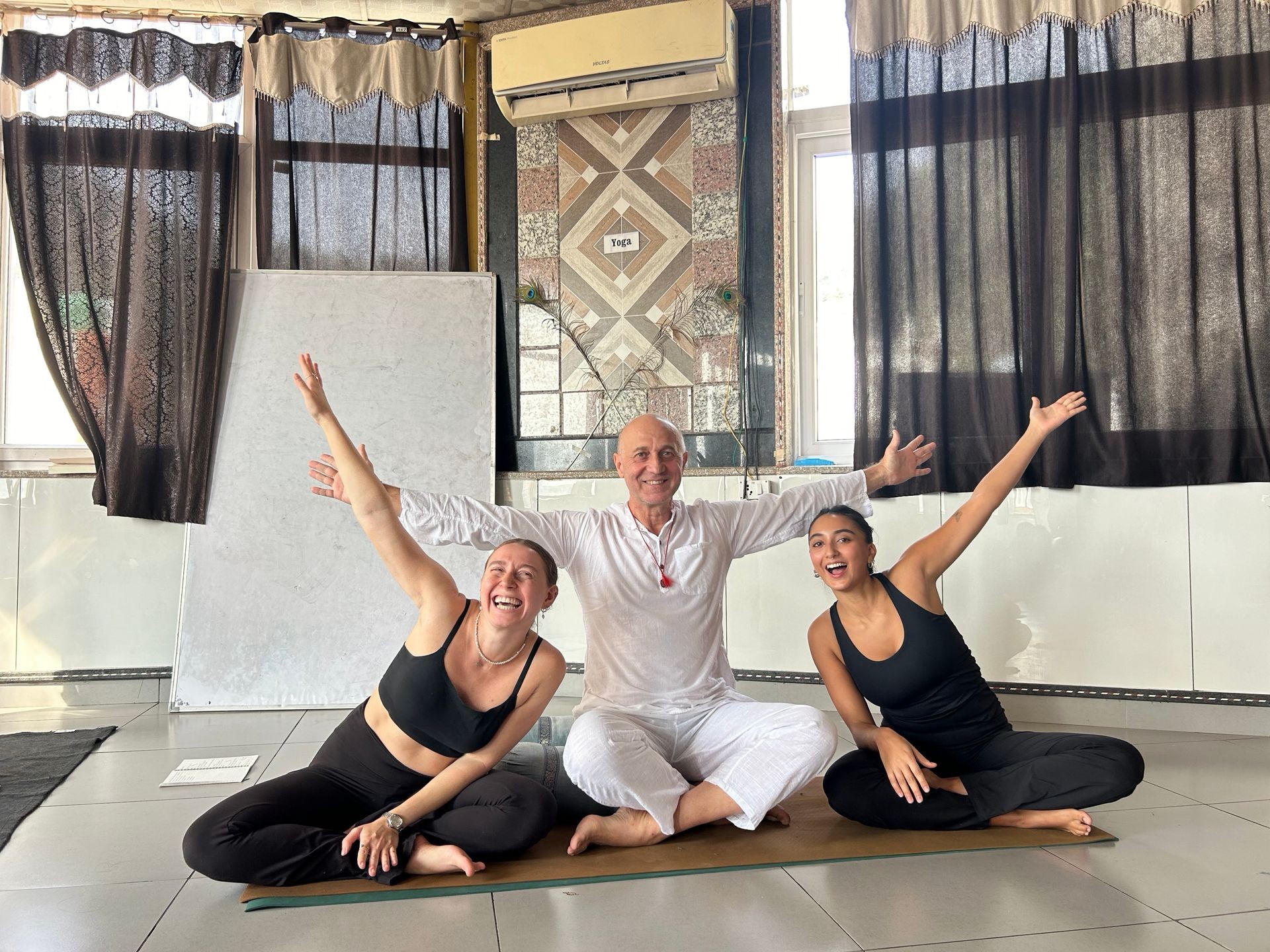 Three people in yoga poses sitting on a mat indoors with arms raised, smiling.